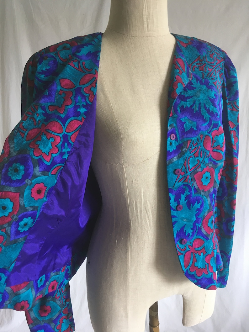 Op de afbeelding: Een paars en turquoise bloemenpatroon jas met een knoopsluiting. De jas is open en de voering is effen paars.