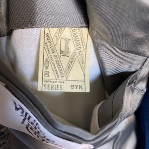May include: A grey garment interior featuring a cream-colored union label with the text Workers of America, Copyright 1969, Series SYK, and a sewing machine icon. A white tag with partial text is visible near the bottom of the grey lining.