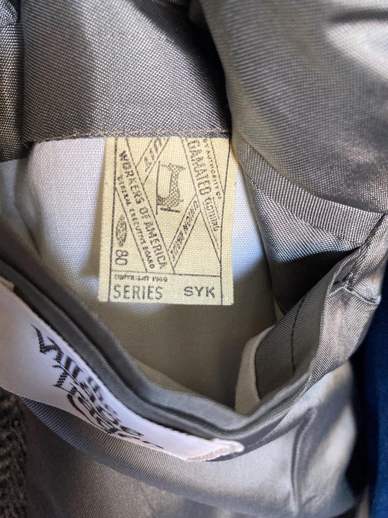 May include: A grey garment interior featuring a cream-colored union label with the text Workers of America, Copyright 1969, Series SYK, and a sewing machine icon. A white tag with partial text is visible near the bottom of the grey lining.