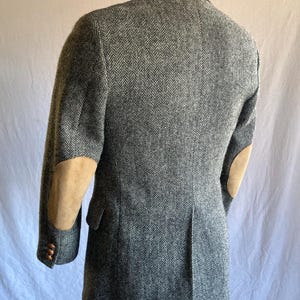 May include: A grey and white herringbone patterned blazer featuring tan suede elbow patches on the sleeves. The jacket includes a notched lapel, a single vent at the back, and brown buttons on the cuffs, shown from a rear-facing angle.