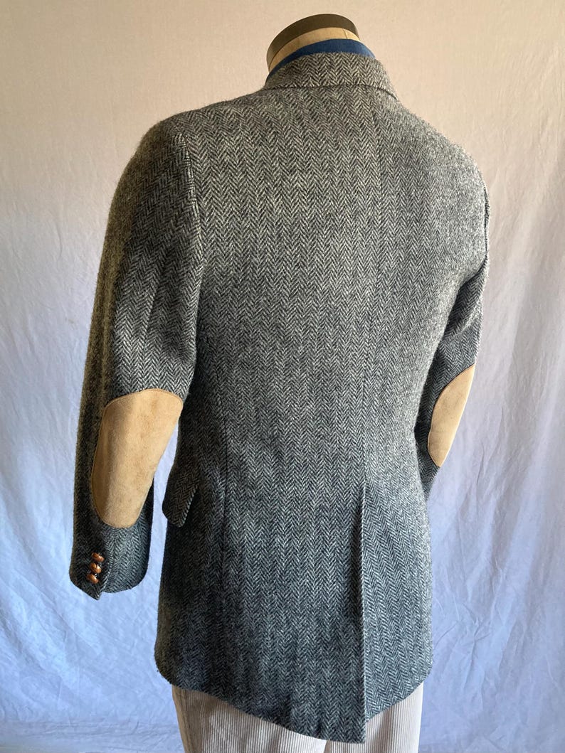 May include: A grey and white herringbone patterned blazer featuring tan suede elbow patches on the sleeves. The jacket includes a notched lapel, a single vent at the back, and brown buttons on the cuffs, shown from a rear-facing angle.