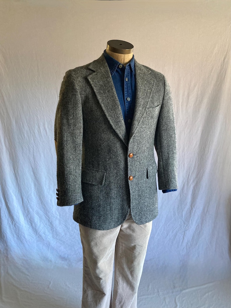 May include: A grey herringbone blazer with two brown buttons, worn over a dark blue button-down shirt and light beige trousers. The blazer features notched lapels, a chest pocket, and two flap pockets on the front, displayed on a mannequin.
