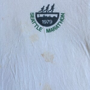 May include: White t-shirt with a green and black logo that says "Seattle Marathon 1979". The logo features a silhouette of runners.