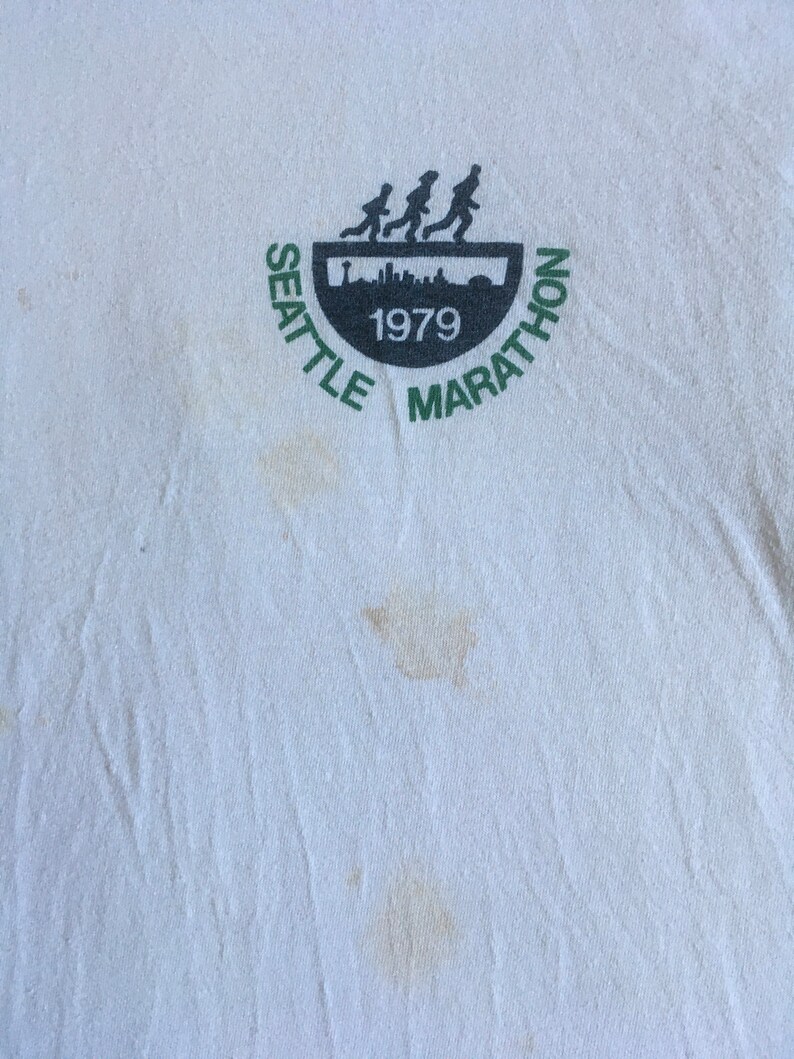 May include: White t-shirt with a green and black logo that says "Seattle Marathon 1979". The logo features a silhouette of runners.