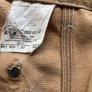 May include: Close-up of brown Carhartt work pants. The tag shows the brand name, care instructions, and sizes 32 and 34. The pants have a button closure and visible stitching.