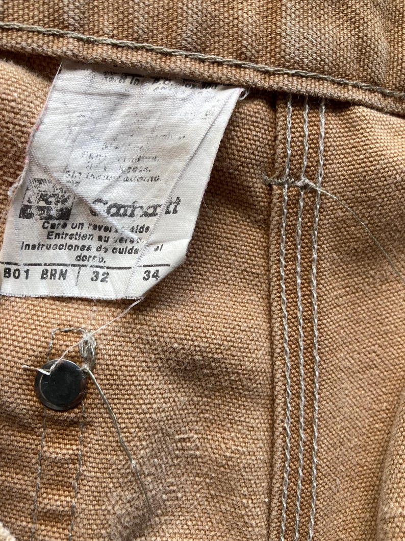 May include: Close-up of brown Carhartt work pants. The tag shows the brand name, care instructions, and sizes 32 and 34. The pants have a button closure and visible stitching.