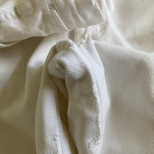 May include: Close-up of a white denim jacket, highlighting the fabric's texture and stitching. The image focuses on the collar, button area, and a folded sleeve, emphasising the garment's construction and design. The off-white colour suggests a vintage or well-worn aesthetic.