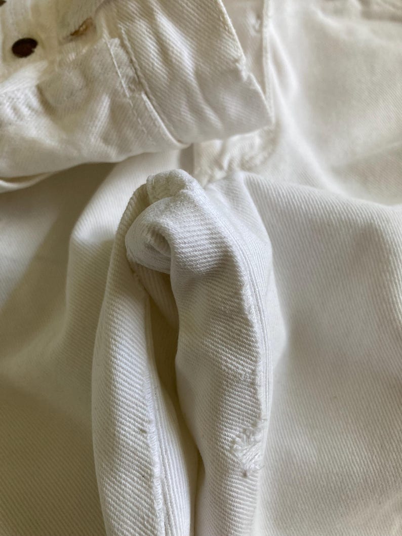 May include: Close-up of a white denim jacket, highlighting the fabric's texture and stitching. The image focuses on the collar, button area, and a folded sleeve, emphasising the garment's construction and design. The off-white colour suggests a vintage or well-worn aesthetic.