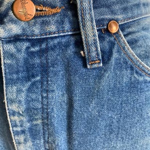 Pu&ograve; includere: Primo piano di jeans blu. I jeans presentano bottoni e rivetti color rame. I jeans hanno un classico design a cinque tasche con chiusura con bottone e cerniera. Il denim ha un aspetto sbiadito e usurato.