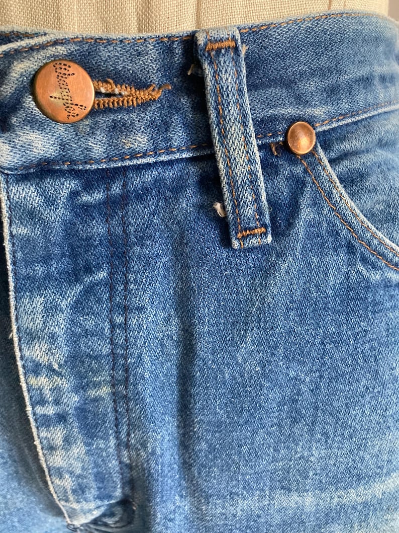 Pu&ograve; includere: Primo piano di jeans blu. I jeans presentano bottoni e rivetti color rame. I jeans hanno un classico design a cinque tasche con chiusura con bottone e cerniera. Il denim ha un aspetto sbiadito e usurato.