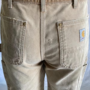 May include: Close-up of tan work pants with multiple pockets and a brown waistband. The pants have a Carhartt logo patch on the back pocket. The pants are made of a durable, heavy-duty fabric.