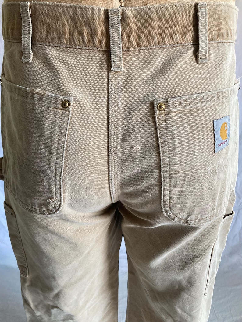 May include: Close-up of tan work pants with multiple pockets and a brown waistband. The pants have a Carhartt logo patch on the back pocket. The pants are made of a durable, heavy-duty fabric.