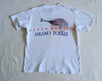 May include: White t-shirt featuring a graphic of a helicopter flying over palm trees and a sunset. The text "Ocean Pacific Island Tours" is printed below the graphic.