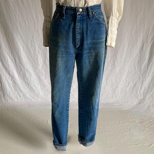 Pu&ograve; includere: Jeans blu in denim a vita alta con chiusura a bottone. I jeans hanno un classico design a cinque tasche e sono risvoltati alle caviglie. Il denim ha un lavaggio sbiadito, che conferisce loro un aspetto vintage. I jeans sono abbinati a una camicetta bianca.
