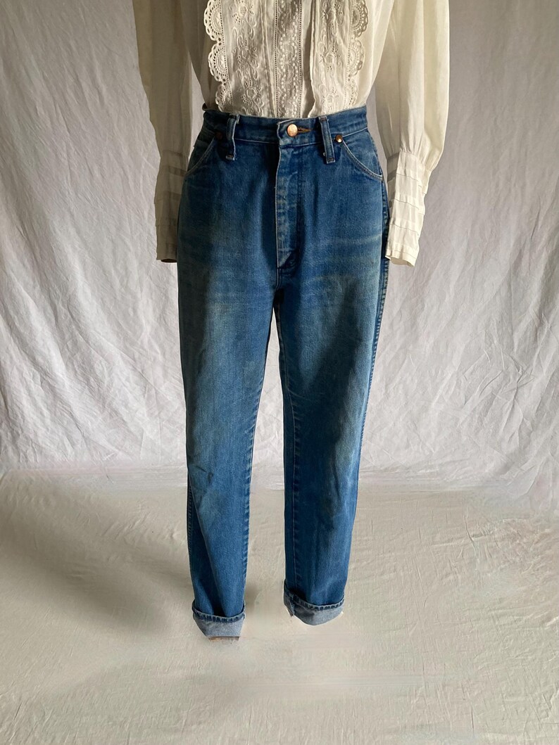 Pu&ograve; includere: Jeans blu in denim a vita alta con chiusura a bottone. I jeans hanno un classico design a cinque tasche e sono risvoltati alle caviglie. Il denim ha un lavaggio sbiadito, che conferisce loro un aspetto vintage. I jeans sono abbinati a una camicetta bianca.