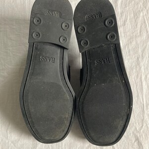 May include: Two black shoe soles are shown, revealing the textured rubber undersides. The soles are branded with "BASS" and have a grip pattern. The shoes appear to be in good condition.