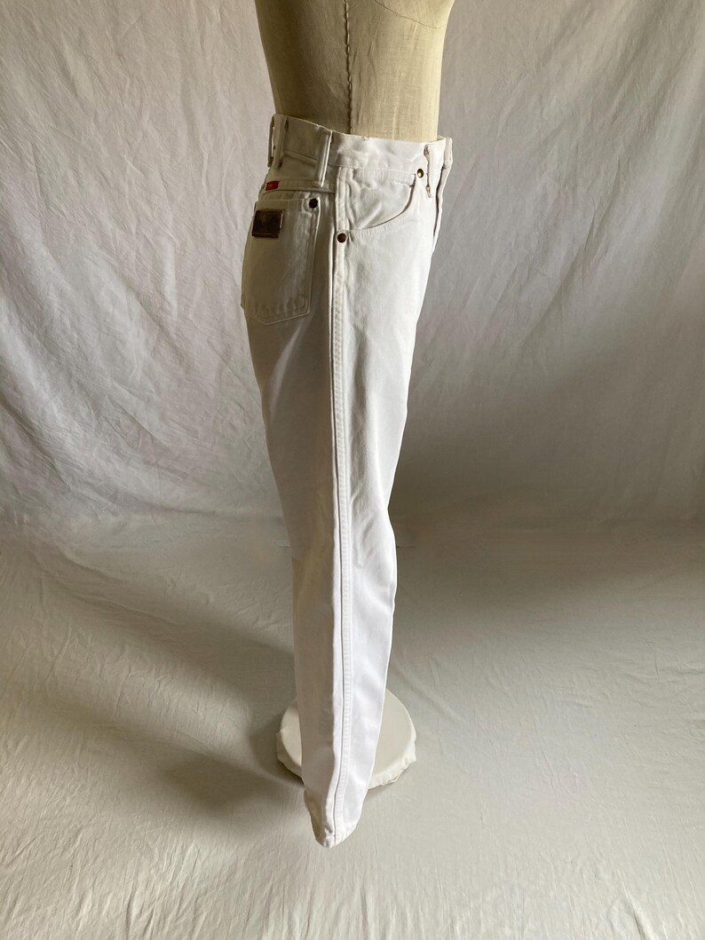 May include: White denim jeans with a classic five-pocket design. The jeans have a straight leg cut and a red logo patch on the back pocket. The jeans are displayed on a mannequin.