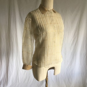 May include: A sheer, ivory-colored blouse with gold stripes and a gold collar. The blouse has a button-up front and long sleeves.