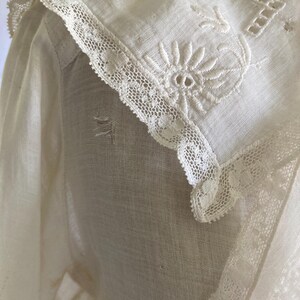 May include: Close-up of a vintage white blouse with intricate floral embroidery and delicate lace trim. The collar features a detailed embroidered flower and geometric patterns. The fabric appears to be a lightweight linen or cotton.