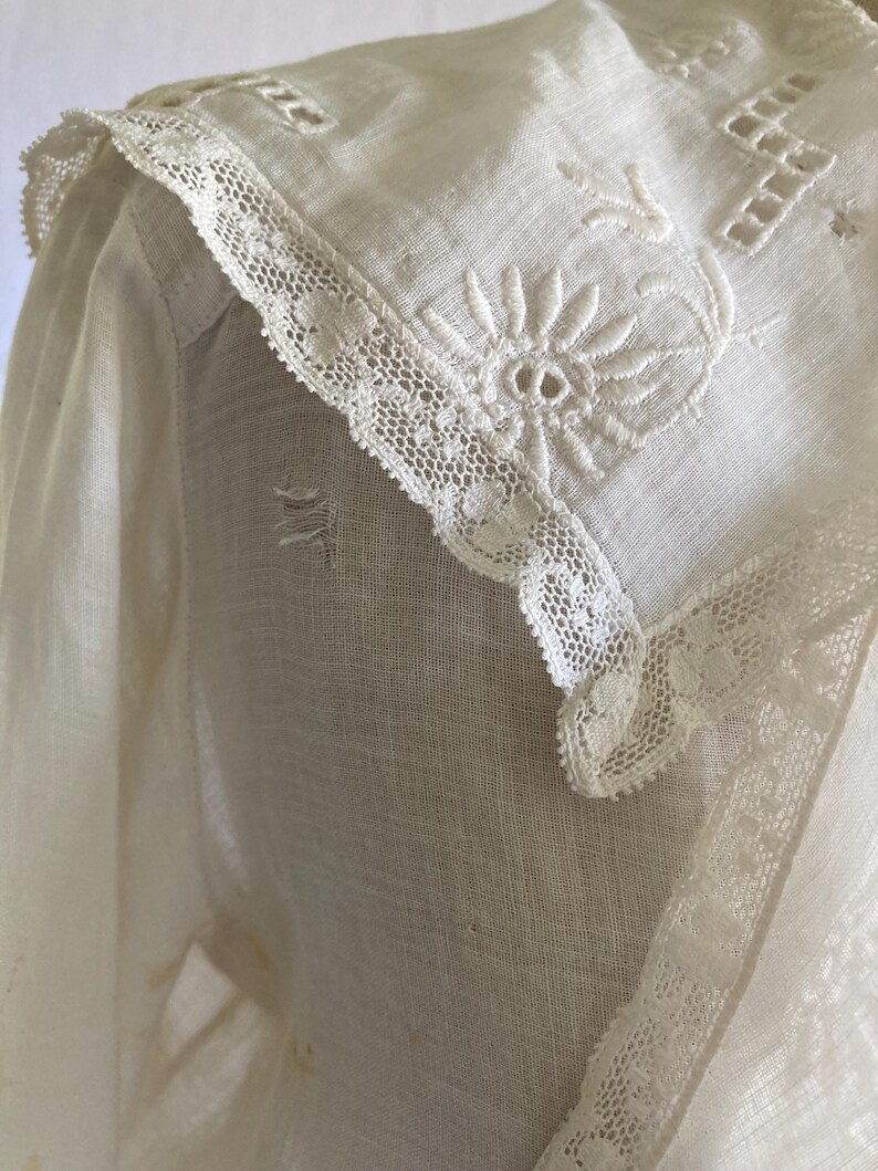 May include: Close-up of a vintage white blouse with intricate floral embroidery and delicate lace trim. The collar features a detailed embroidered flower and geometric patterns. The fabric appears to be a lightweight linen or cotton.