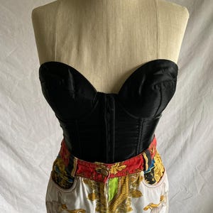 May include: A black satin strapless corset with boning and a fitted silhouette. The corset is paired with white denim jeans featuring a colorful, patterned waistband and a white, gold, and blue print.
