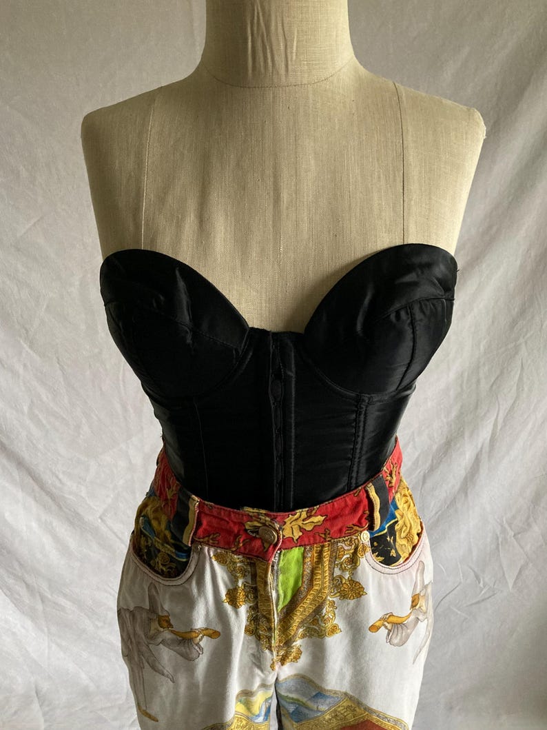 May include: A black satin strapless corset with boning and a fitted silhouette. The corset is paired with white denim jeans featuring a colorful, patterned waistband and a white, gold, and blue print.