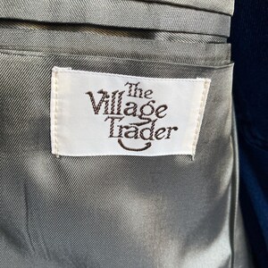 May include: A grey satin-like lining inside a garment featuring a rectangular white fabric label stitched onto the material. The label displays the text The Village Trader in dark brown lettering with a curved line underneath the word Trader.
