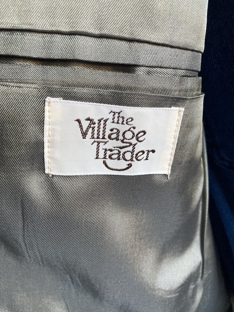 May include: A grey satin-like lining inside a garment featuring a rectangular white fabric label stitched onto the material. The label displays the text The Village Trader in dark brown lettering with a curved line underneath the word Trader.