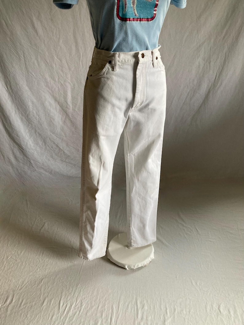 May include: White denim jeans with a classic five-pocket design. The jeans have a straight leg cut and a button and zip closure. The jeans are displayed on a mannequin.