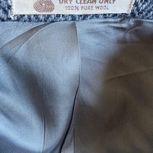 May include: A gray herringbone wool garment interior featuring a smooth, light gray lining. A rectangular fabric label is stitched at the top, displaying a yarn icon and the text: DRY CLEAN ONLY 100% PURE WOOL.