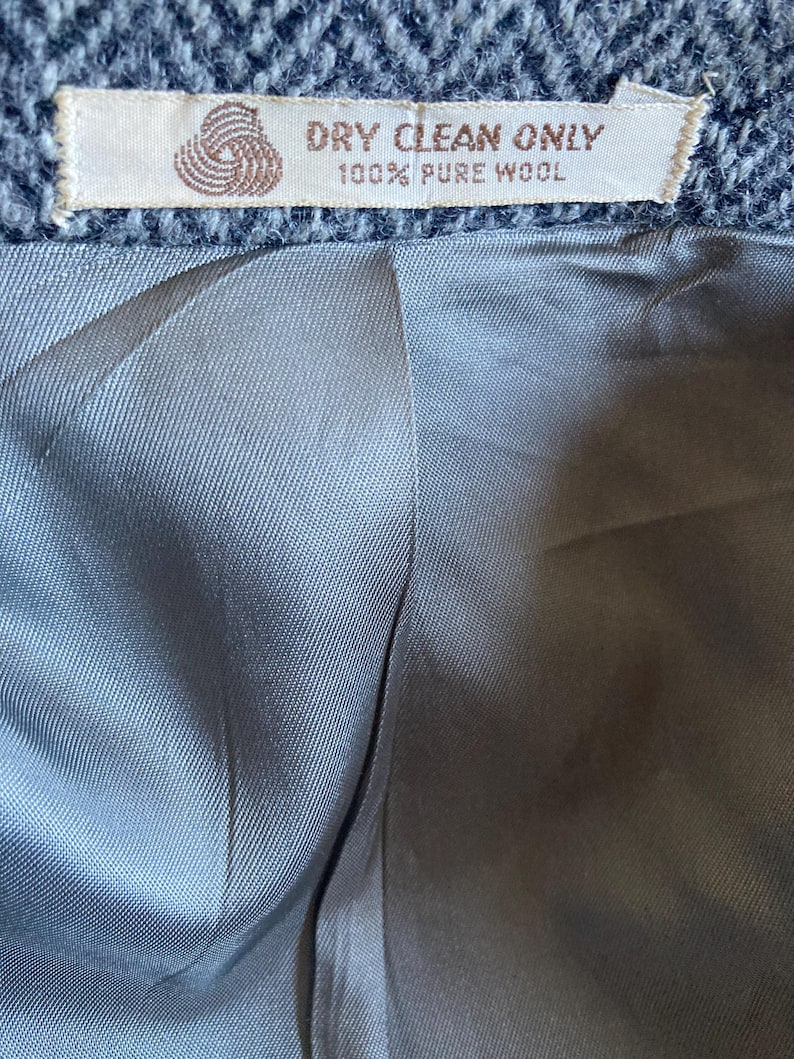 May include: A gray herringbone wool garment interior featuring a smooth, light gray lining. A rectangular fabric label is stitched at the top, displaying a yarn icon and the text: DRY CLEAN ONLY 100% PURE WOOL.