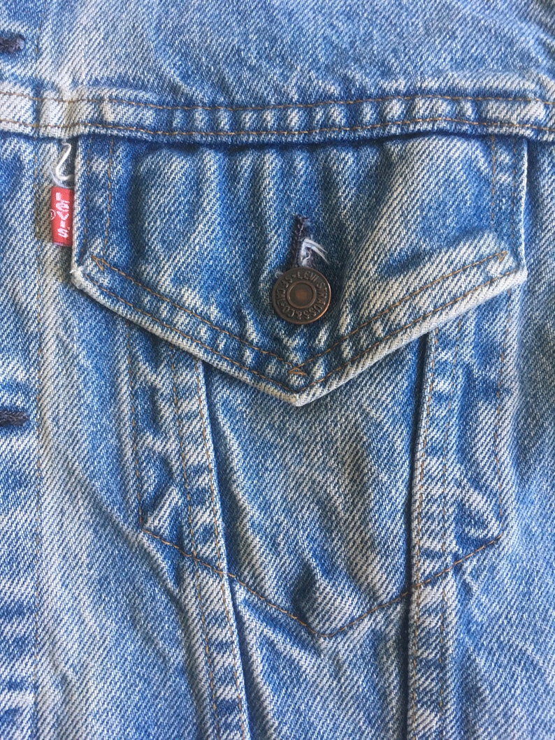 May include: Close-up of a blue denim jacket with a pocket and a metal button. The pocket has a small Levi's label.