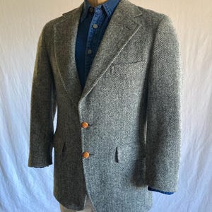May include: A grey and white herringbone patterned blazer featuring notched lapels, two brown buttons at the front closure, and a single chest pocket. The blazer is layered over a blue button-down shirt and light-colored trousers on a mannequin.