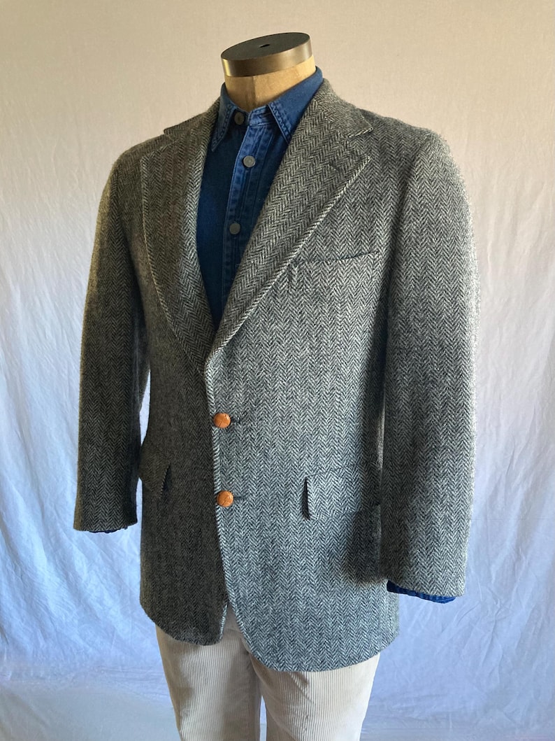 May include: A grey and white herringbone patterned blazer featuring notched lapels, two brown buttons at the front closure, and a single chest pocket. The blazer is layered over a blue button-down shirt and light-colored trousers on a mannequin.