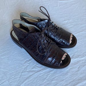 May include: A pair of brown leather open-toe sandals with a crocodile texture. The sandals have laces and a slingback design. The open toe area has a pink and white floral pattern. The shoes are on a white surface.