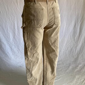 May include: Tan work pants with multiple pockets and a wide belt loop. The pants are made of a durable fabric and have a relaxed fit. The back features two large pockets and a small logo patch.