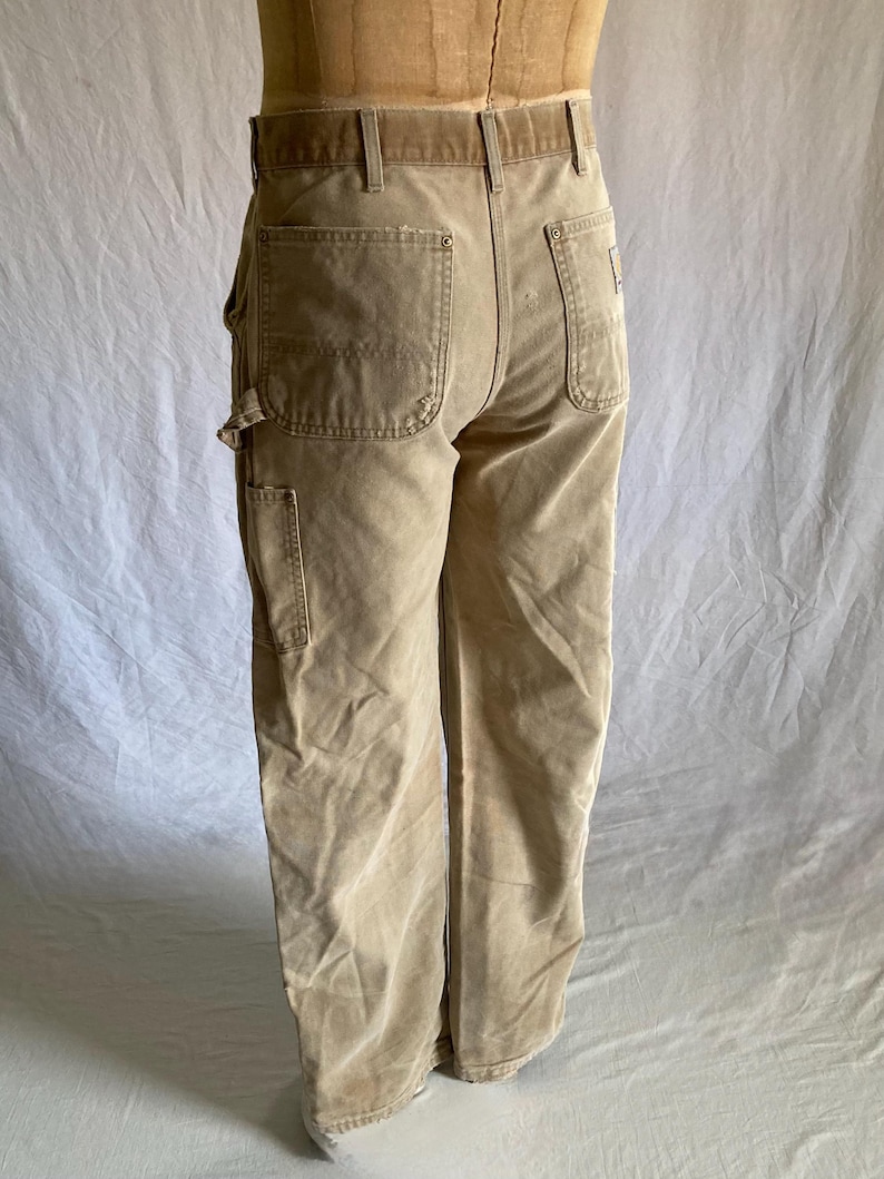 May include: Tan work pants with multiple pockets and a wide belt loop. The pants are made of a durable fabric and have a relaxed fit. The back features two large pockets and a small logo patch.
