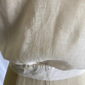 May include: Close-up of a cream-colored, vintage blouse. The fabric appears to be lightweight and slightly sheer, with subtle texture and a gathered waistline. The blouse is displayed on a mannequin, showcasing its shape and design.