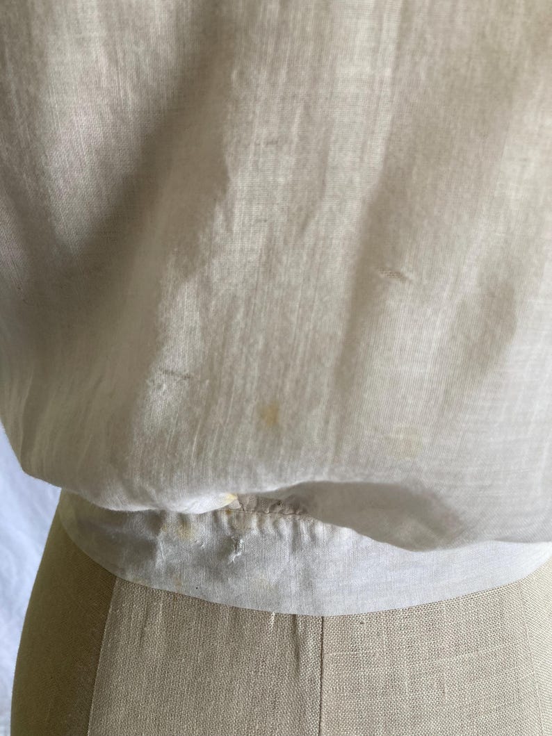 May include: Close-up of a cream-colored, vintage blouse. The fabric appears to be lightweight and slightly sheer, with subtle texture and a gathered waistline. The blouse is displayed on a mannequin, showcasing its shape and design.