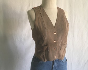 May include: A brown suede vest with a V-neck and button closure. The vest has two pockets on the front and is worn over a pair of blue jeans.