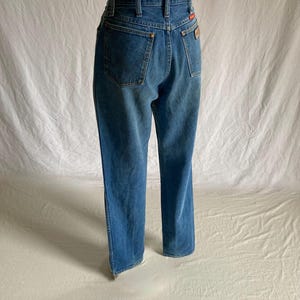 Pu&ograve; includere: Jeans in denim blu con vita alta e gamba dritta. I jeans hanno due tasche posteriori, passanti per la cintura e una piccola toppa rossa e marrone sul retro. I jeans sono esposti su un manichino.