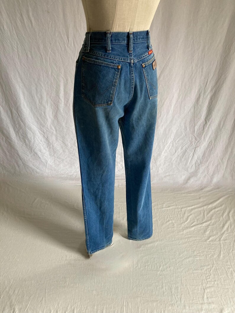 Pu&ograve; includere: Jeans in denim blu con vita alta e gamba dritta. I jeans hanno due tasche posteriori, passanti per la cintura e una piccola toppa rossa e marrone sul retro. I jeans sono esposti su un manichino.