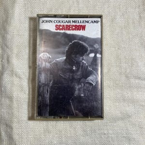 May include: A vintage cassette tape of John Cougar Mellencamp's album "Scarecrow." The album cover features a black and white image of a man leaning on a wooden fence. The album title is in red.