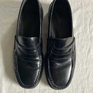 May include: A pair of black leather penny loafers. These shoes feature a classic design with a rounded toe, a strap across the vamp, and a stitched sole. The leather has a glossy finish.