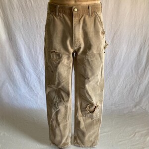 May include: Tan work pants with visible wear and tear, including patches and holes. The pants have a button closure and multiple pockets. The fabric appears to be a durable, heavy-duty material, suggesting they are designed for work or rugged use.