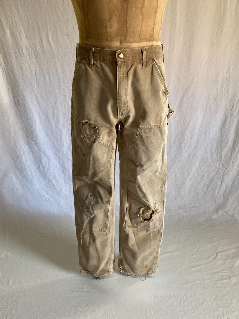May include: Tan work pants with visible wear and tear, including patches and holes. The pants have a button closure and multiple pockets. The fabric appears to be a durable, heavy-duty material, suggesting they are designed for work or rugged use.