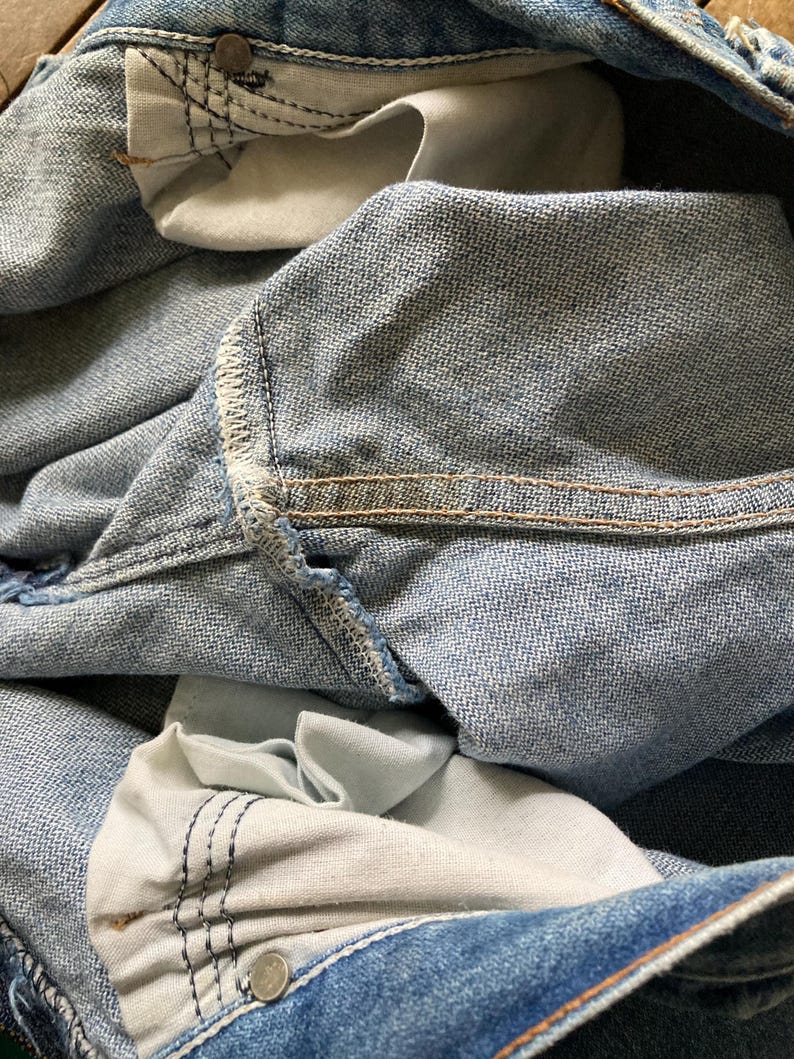 Pu&ograve; includere: Primo piano di jeans in denim blu chiaro, che mostra le tasche interne e i dettagli delle cuciture. Le tasche sono di una tonalit&agrave; di blu pi&ugrave; chiara e le cuciture sono rinforzate con filo a contrasto. Rivetti e bottoni metallici visibili.