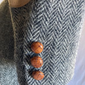May include: A gray and white herringbone patterned blazer sleeve featuring a tan suede elbow patch and three round, brown, woven leather buttons aligned vertically along the cuff of the garment sleeve.
