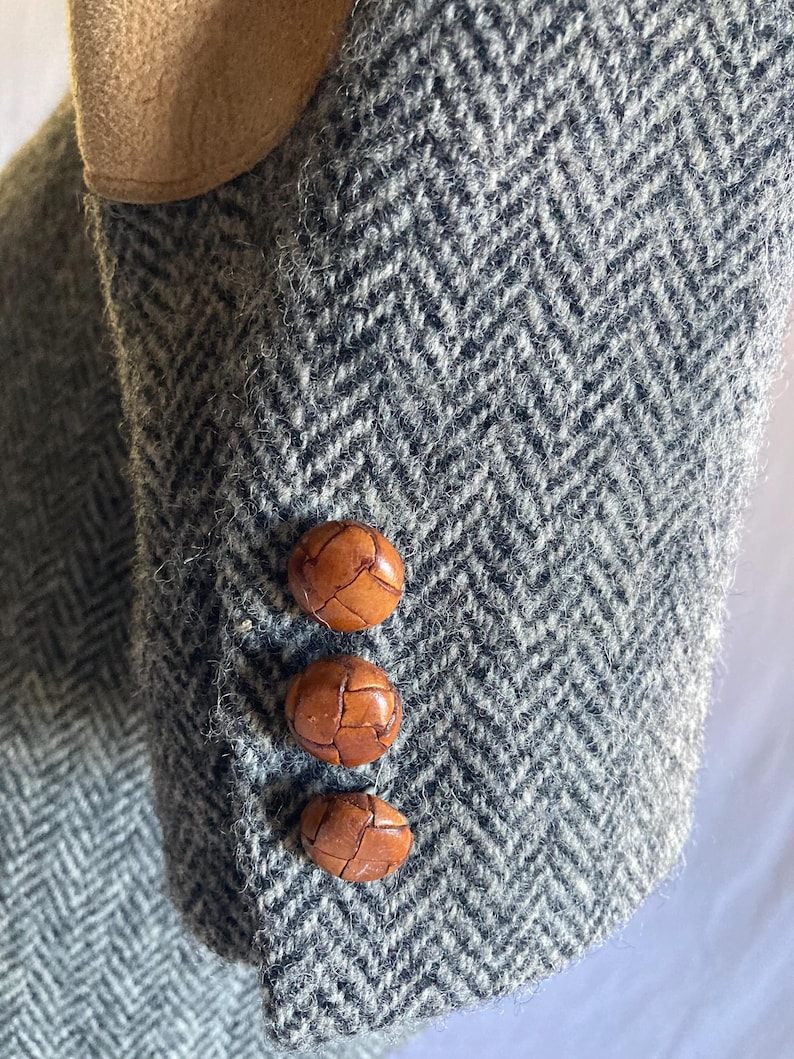 May include: A gray and white herringbone patterned blazer sleeve featuring a tan suede elbow patch and three round, brown, woven leather buttons aligned vertically along the cuff of the garment sleeve.