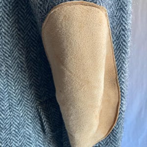 May include: A gray and white herringbone patterned blazer sleeve featuring a large, oval-shaped tan suede elbow patch stitched onto the fabric. The sleeve is textured with a classic woven pattern and includes a visible brown button at the cuff.